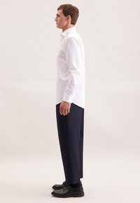 Man in profiel staand, gekleed in een wit overhemd, marineblauwe broek, zwarte sokken en zwarte nette schoenen tegen een neutrale achtergrond.