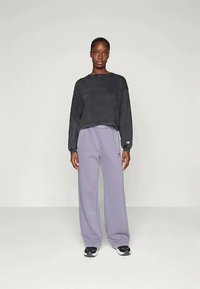 Ženska stoji oblečena v črno kratko majico, ohlapne svetlo vijolične trenirke in črno-bele superge na enotnem svetlem ozadju.