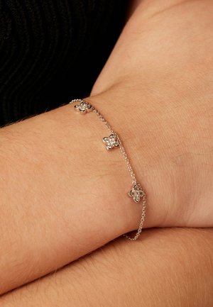 Zilveren kettingarmband met drie bloemetjesvormige bedels met heldere edelstenen, rustend op een lichte huidkleurige pols.