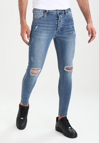 Man som bär ljusblå slitna skinny jeans och svarta sneakers, står mot en enfärgad ljus bakgrund.