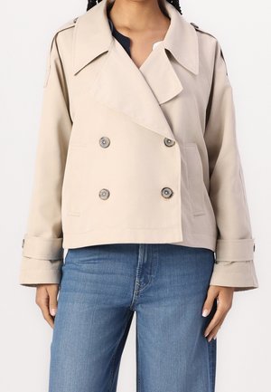 Vrouw die een beige, dubbelgeknoopte, korte trenchcoat draagt met brede revers en omslagmouwen, gecombineerd met blauwe spijkerbroeken.