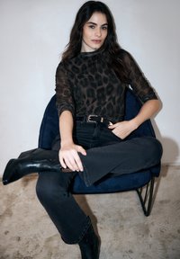 Zwarte doorschijnende top met luipaardprint gecombineerd met zwarte high-waisted jeans, zittend op een gestructureerde blauwe stoel met metalen poten, met enkellaarzen.