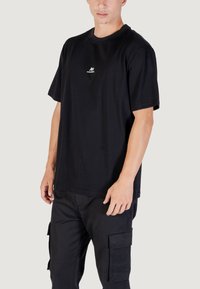 Camiseta de algodón negra con mangas cortas, que presenta un pequeño logo blanco en el pecho y un ajuste relajado. Combinada con pantalones cargo negros.