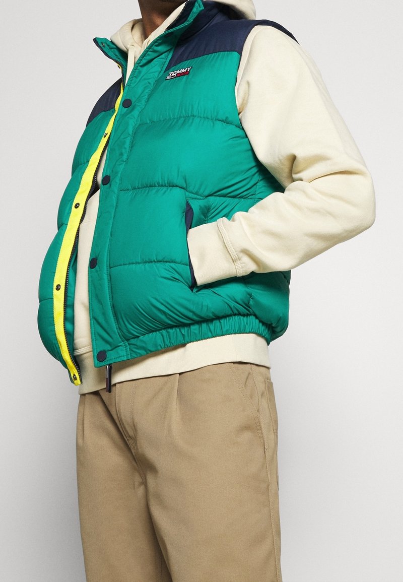 Chaleco acolchado colorido en verde con hombros azul marino, que presenta una cremallera amarilla, dos bolsillos laterales y dobladillo elástico, llevado sobre una sudadera con capucha clara.