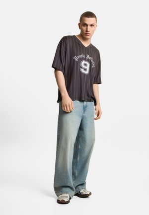 Młody mężczyzna stojący w jasnych, szerokich dżinsach, czarnej pasiakowanej koszulce z napisem „Prosto Infinity” i numerem 9 oraz białych butach sportowych na białym tle.