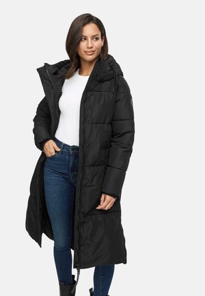 Woman wearing a long black quilted coat with hood, white shirt, blue jeans, and black boots, standing with one hand in pocket.