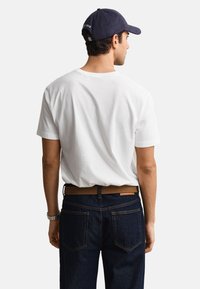 Mann trägt ein weißes kurzärmeliges T-Shirt, dunkelblaue Jeans mit braunem Gürtel, eine schwarze Uhr und eine dunkelblaue Kappe, blickt weg.