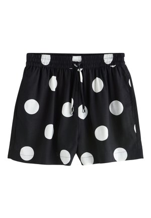 Sorte shorts med store hvide prikker båret af en person, der løfter en matchende sort top med hvide prikker.