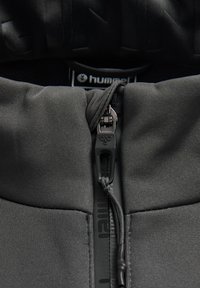 Sort zip-up hoodie med et glat, tekstureret stof, der har en høj krave, lynlåslukning foran og et logo-mærke. Diskrete syninger er synlige.