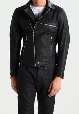 Homme portant une veste en cuir noire ajustée avec une fermeture éclair asymétrique et des poches zippées sur une chemise blanche et un pantalon noir.