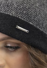 Gorro de punto en tonos negro y gris, con una etiqueta circular plateada que dice "vivisence". La textura parece suave y cálida.
