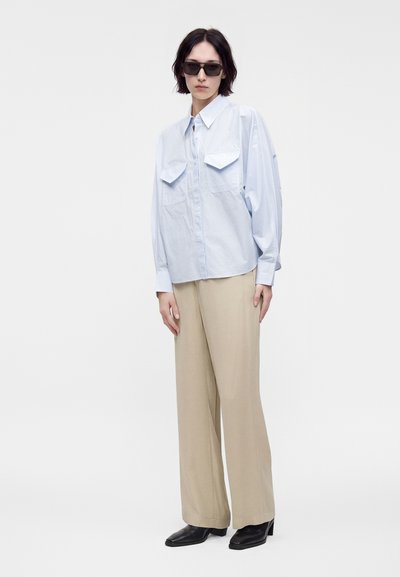 Femme portant une chemise oversize bleu clair à boutons, un pantalon beige à jambes larges, des chaussures noires à talons et des lunettes de soleil foncées, debout devant un fond blanc.