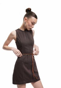 Bruine katoenen jurk met een aansluitende bodice, mouwloos ontwerp, rechte rok en een beige riem die gestrikt kan worden. Heeft zijzakken en stikseldetails.