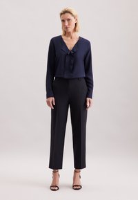 Nainen seisoo yksiväristä taustaa vasten pukeutuneena tummansiniseen pitkähihaiseen puseroon, mustiin räätälöityihin housuihin ja mustiin avokkaisiin.