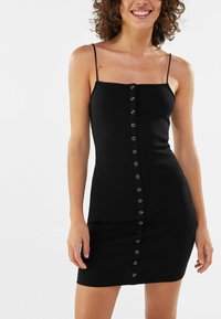 Svart ärmlös klänning med en figurnära passform, tillverkad av ett elastiskt tyg. Har en knappdetalj fram med silverfärgad hårdvara och en mjuk textur.
