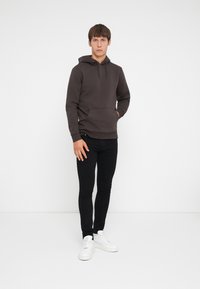Dark brown hoodie with a kangaroo pocket, drawstring hood, worn with black skinny jeans and white sneakers. Plain background.
