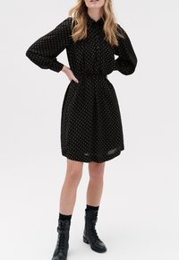 Zwart jurkje met witte stippen, lange mouwen, een knoopsluiting aan de voorkant, een getailleerde taille en een A-lijn rok; gecombineerd met zwarte veterschoenen.