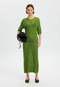 Vestido verde de punto con una forma larga y recta y patrones geométricos, combinado con un bolso negro de tejido y zapatos planos negros.