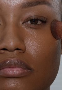 Nahaufnahme eines Gesichts mit glatter, bronzierter Haut, neutralen Lippen und einem sichtbaren Auge. Ein Makeup-Pinsel berührt sanft die Wange und verblendet das Produkt.