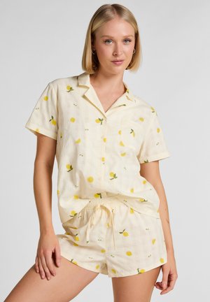 Frau trägt hellgelbes Kurzarmhemd und passende Shorts mit kleinen Zitronenmustern, posiert mit einer Hand auf dem Oberschenkel.