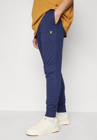 Tmavě modré tepláky s elastickými manžetami, bočními kapsami a malým žlutým logem na stehně, vyrobené z měkké a pohodlné tkaniny.