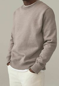 Sudadera marrón claro con cuello redondo y puños acanalados, textura suave y dobladillo recto. Lleva puesta una camiseta blanca y pantalones claros.