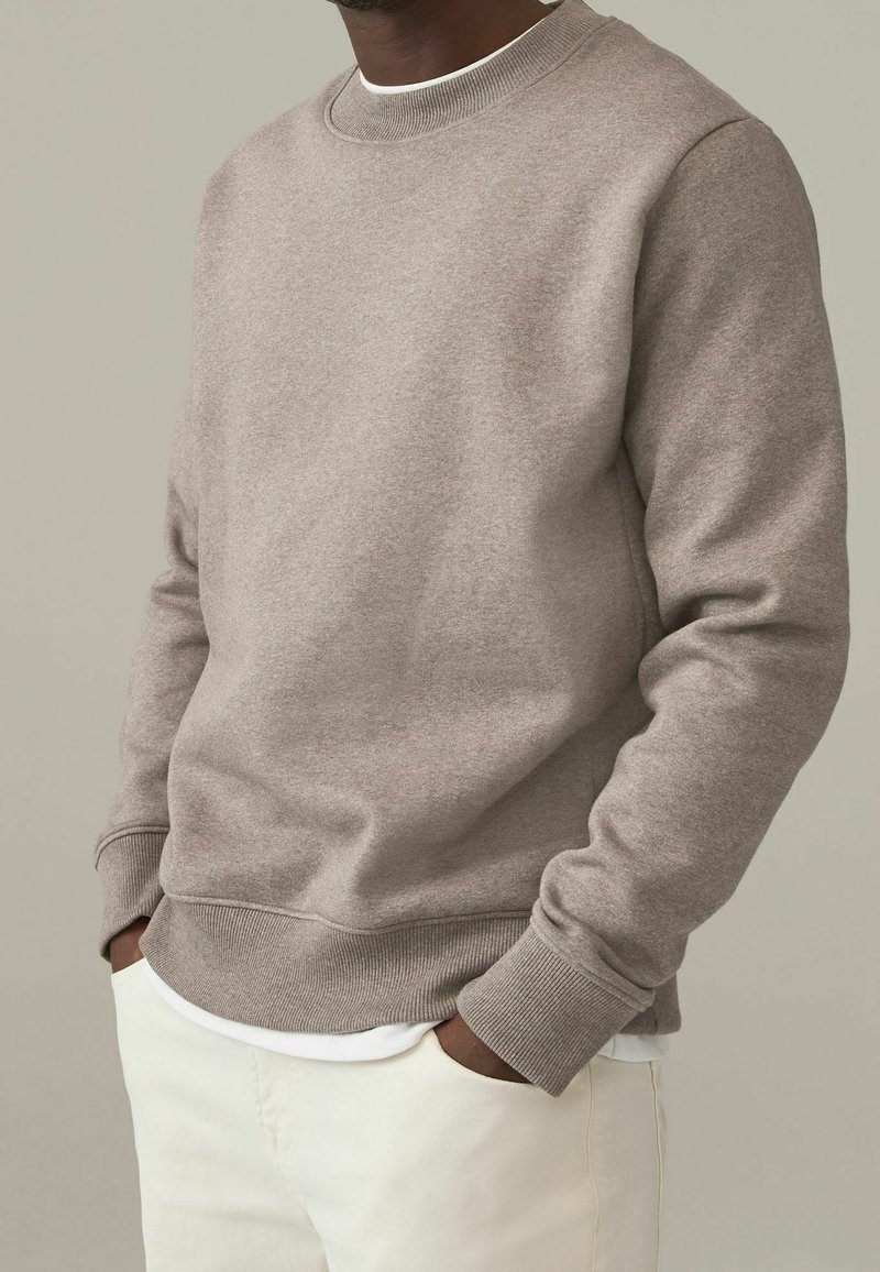 Sudadera marrón claro con cuello redondo y puños acanalados, textura suave y dobladillo recto. Lleva puesta una camiseta blanca y pantalones claros.