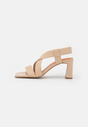 Beige sandal med åbne tæer, krydsede remme, slingback og en medium blokhæl på en ensfarvet hvid baggrund.