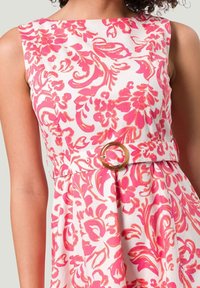 Blumenkleid mit einem pink-weißen Design, ärmelloser Schnitt und einem Taillengürtel mit einem runden goldfarbenen Ringdetail. Aus Baumwollmaterial.