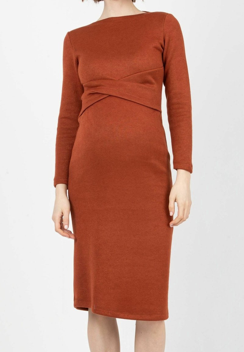 Femme portant une robe couleur rouille à manches longues, longueur genou, avec un détail torsadé à la taille.
