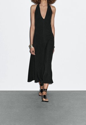 Black halter jumpsuit with a deep V-neck and wide legs, made from a lightweight fabric. Paired with black strappy sandals.