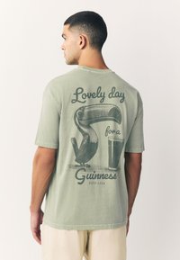 Zelena bombažna majica z velikim motivom tukana in kozarca ter napisom "Čudovit dan za Guinness" v vintidž slogu.