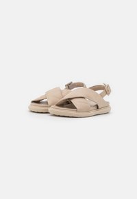Beige sandaalit, joissa on tikattu muotoilu, ristisiteet ja säädettävä nilkkahihna, kevyellä ja teksturoidulla pohjalla.