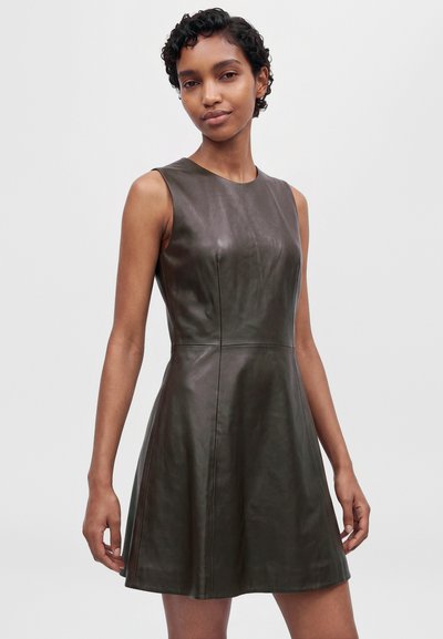 Un mannequin pose sur un fond uni, vêtu d’une robe sans manches vert olive foncé, d’aspect cuir, avec un corsage ajusté et une jupe évasée.
