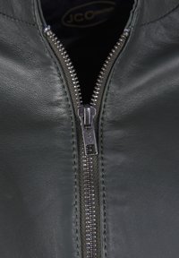 Grön läderjacka med en silverdragkedja, med synliga sömmar och en slät yta. Fodret är svart med en logotypetikett inuti.