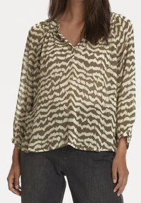 Blouse ample et transparente de couleur vert olive avec un motif abstrait crème, col plissé, fermeture par boutons, et manches longues. Portée avec un jean foncé.