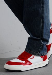 Red and white leather sneaker with textured panels and red laces, paired with dark blue denim jeans. Flat sole with branding detail.