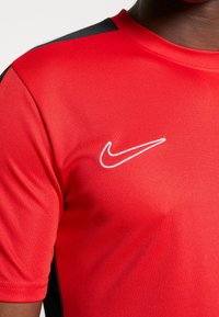 Camisa deportiva roja con paneles de hombro negros y logo de Nike blanco en el lado superior izquierdo del pecho, llevada por una persona de piel oscura.