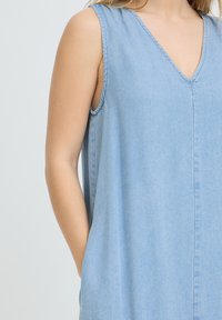 Robe en denim bleu clair sans manches avec col en V portée par une personne, un bras reposant le long du corps, sur un fond uni.