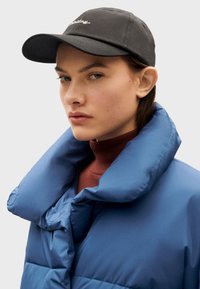 Chaqueta acolchada azul con un alto cuello, detalles de botones y un cuello de tortuga burdeos debajo, complementada con una gorra de béisbol negra.