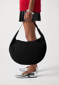 Borsa hobo nera imbottita con silhouette curva e tracolla regolabile, tenuta da una persona che indossa un braccialetto d'argento e scarpe metalliche.