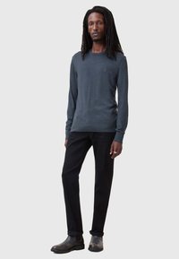 Donkergrijze lange mouwtrui met een ronde hals, gecombineerd met zwarte broek en zwarte leren enkellaarsjes. Model staat tegen een lichte achtergrond.