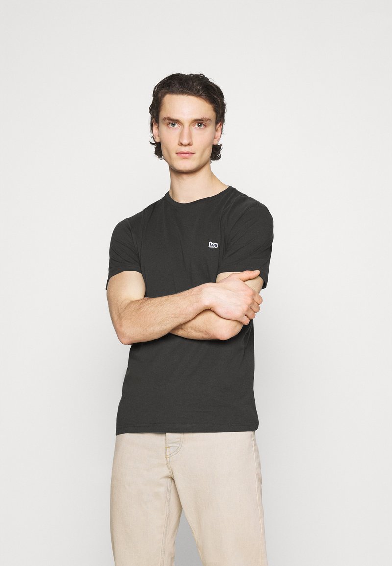 Schwarzes Baumwoll-T-Shirt mit Rundhalsausschnitt und kurzen Ärmeln, mit einem kleinen weißen Logo auf der linken Brust. Model trägt hellfarbige Hosen.