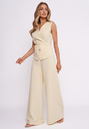 Kobieta z długimi włosami, ubrana w kremowy, bezrękawnikowy top typu wrap z paskiem oraz szerokie spodnie, stojąca na tle jednolitego białego tła.