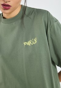 Grøn bomulds t-shirt med en afslappet pasform, der har en gul grafisk tekst "PARIS" foran. Minimalistisk design og blød tekstur.