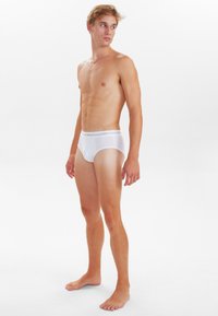 Hvide briefs med en glat tekstur, elastisk talje med logo. Modellen står barfodet og viser et sidesyn.