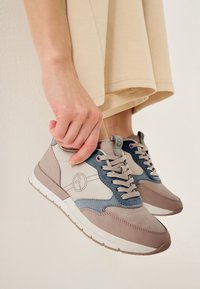 Baskets avec une tige en cuir beige et gris, accents en daim bleu, bout rond, lacets gris et semelle en caoutchouc blanche. Main tenant la chaussure.