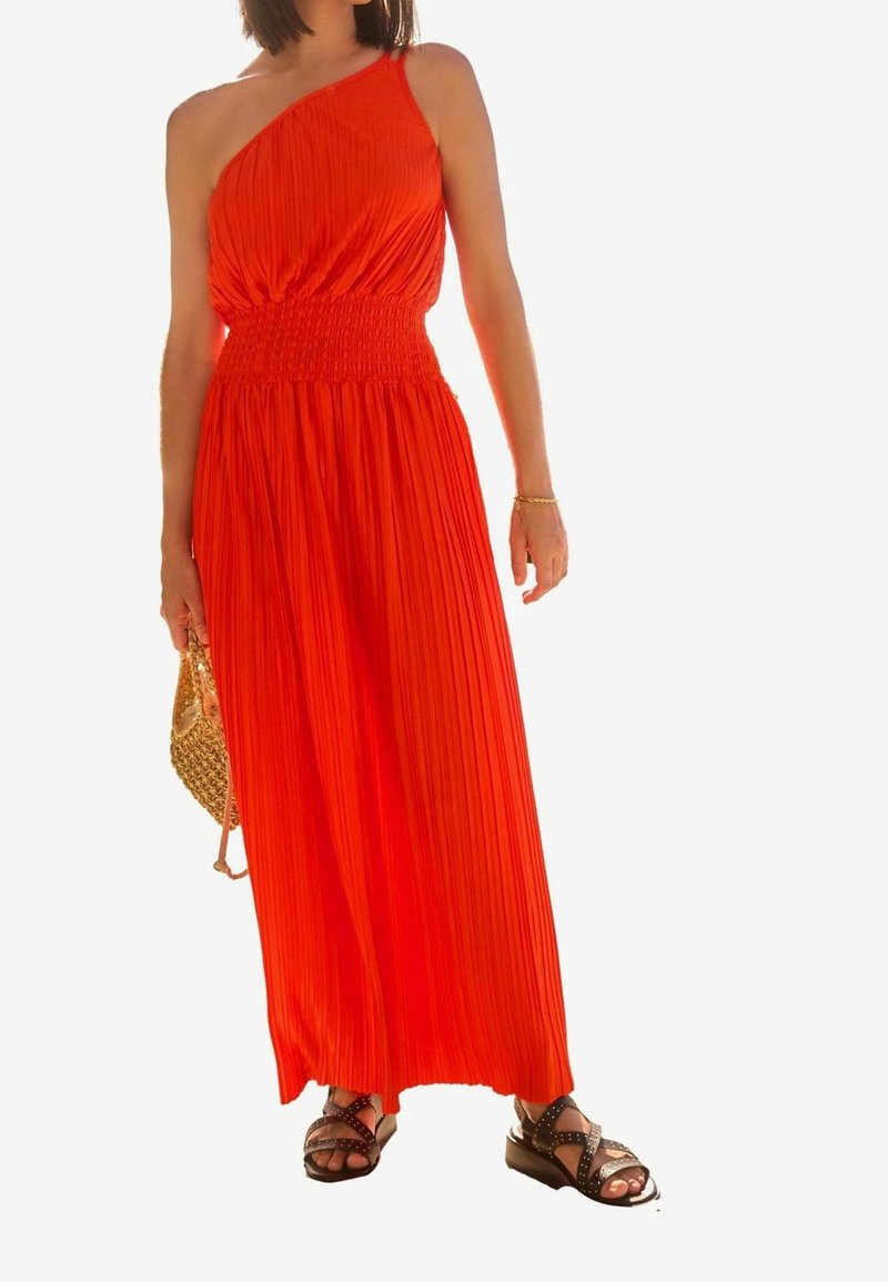 Femme portant une robe longue plissée orange vif à une épaule, des sandales noires cloutées et tenant un petit sac à main tressé.