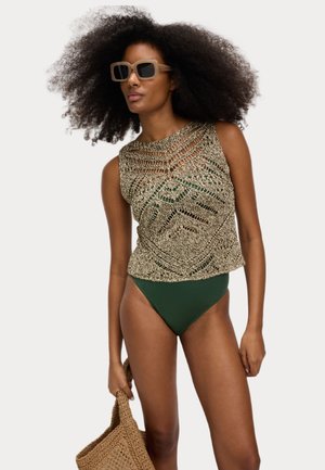 Top de ganchillo en oro y parte inferior de traje de baño verde. Patrón texturizado con detalles intrincados. La modelo sostiene un bolso de mimbre marrón y lleva gafas de sol.