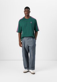 Zielona oversize'owa koszulka z różowymi paskami i logo, zestawiona z szarymi szerokimi spodniami i białymi sneakersami. Minimalistyczny design.